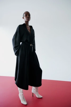 An adult female model in a black dress and white boots poses in a studio with a white and red backdrop for a fashion editorial.