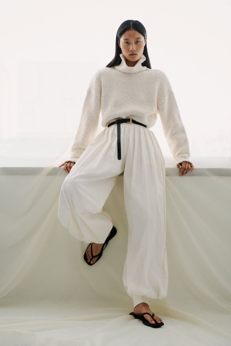 A woman models a relaxed fashion outfit featuring a cream turtleneck sweater, white balloon pants, and black sandals in a bright indoor setting.