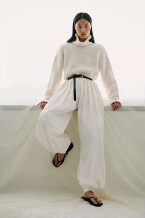 A woman models a relaxed fashion outfit featuring a cream turtleneck sweater, white balloon pants, and black sandals in a bright indoor setting.