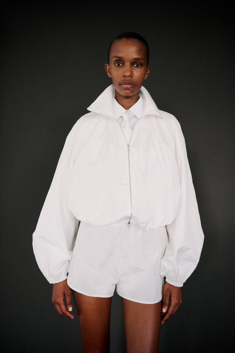 Portrait of a black female model in a white jacket and shorts against a dark gray background in a studio fashion shot.