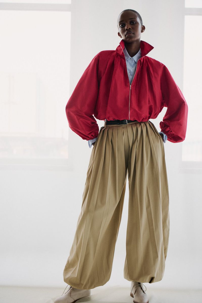 Fashionable adult posing in a red jacket, light blue shirt, and khaki pleated balloon pants against a white background indoors.