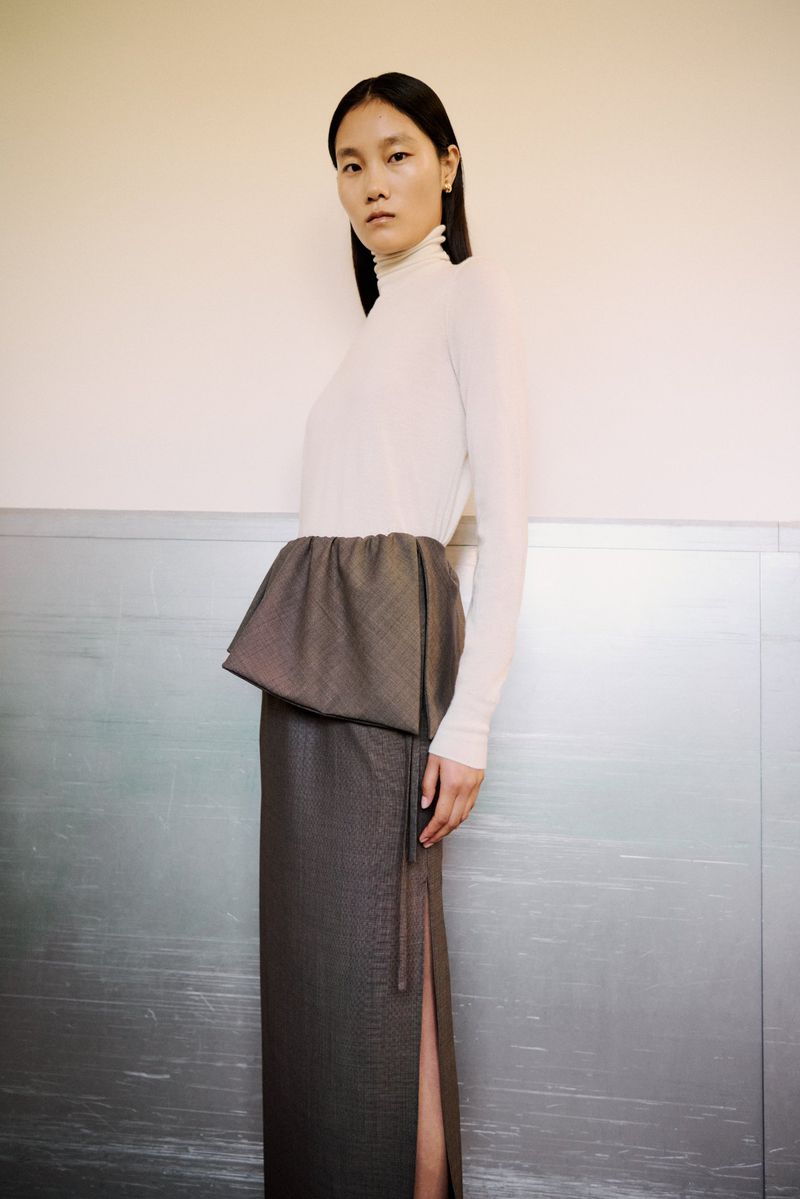 Woman in a long grey skirt with a slit and a white long-sleeve top, posing in front of a silver wall. Fashion editorial shot.