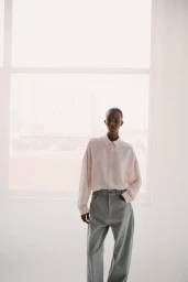 Model Ornella Umutoni posing in a pale pink button-up shirt and grey wide-leg trousers in front of a bright window.