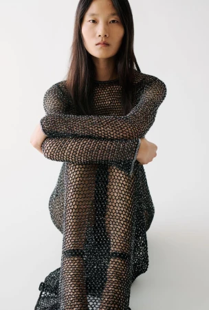 Model Canlan Wang seated in a black open-knit mesh dress against a clean white studio background for Adam Lippes.
