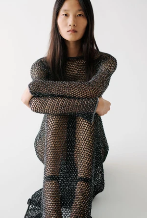 Model Canlan Wang seated in a black open-knit mesh dress against a clean white studio background for Adam Lippes.
