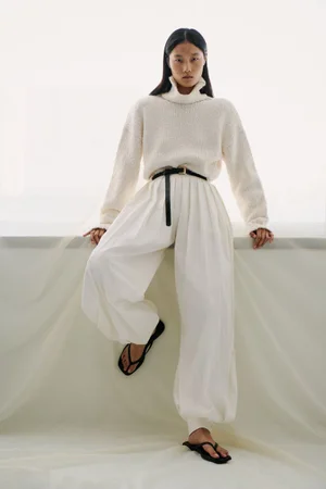 Model Canlan Wang poses in an off-white knit turtleneck sweater and voluminous pleated white trousers for Adam Lippes.