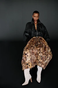 Ornella Umutoni seated in a black zip jacket, voluminous patterned skirt, and cream heeled boots against a dark background.