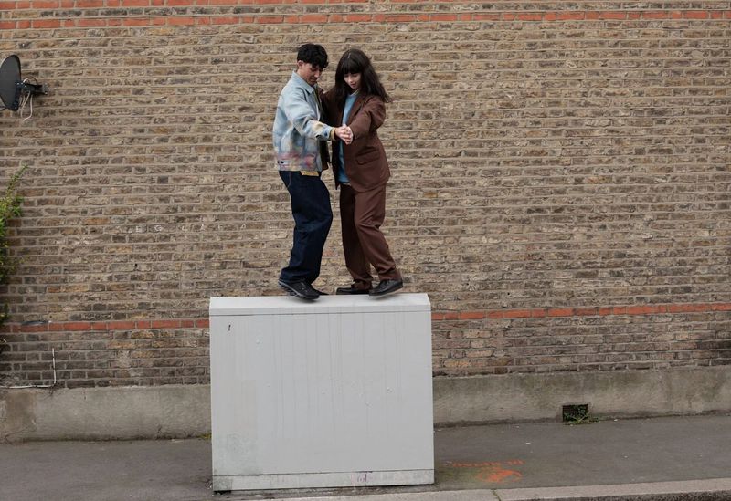 Two adults pose together on a utility box outdoors in casual wear with a brick wall backdrop, captured in a medium shot.