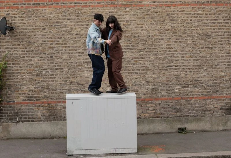 Two adults pose together on a utility box outdoors in casual wear with a brick wall backdrop, captured in a medium shot.