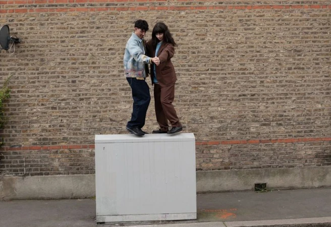 Two adults pose together on a utility box outdoors in casual wear with a brick wall backdrop, captured in a medium shot.