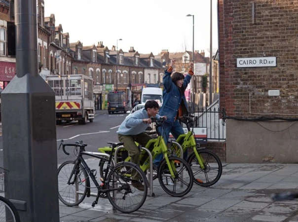 Two young adults ride shared bikes on a city street near buildings, with one raising his arms in celebration, under an overcast sky.