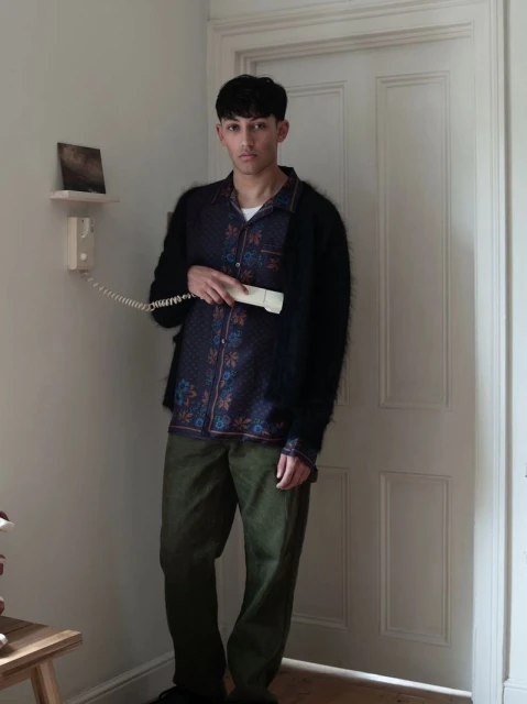 A young adult male stands indoors, holding a telephone receiver, wearing a patterned shirt and dark sweater, with a neutral expression.