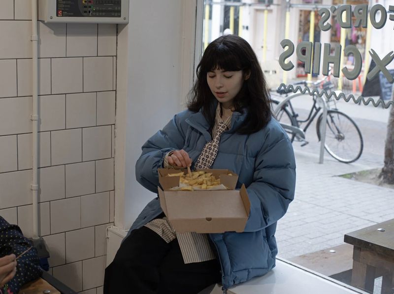 Young woman with dark hair sits indoors eating chips from a cardboard container, wearing a blue puffer jacket and patterned tie.