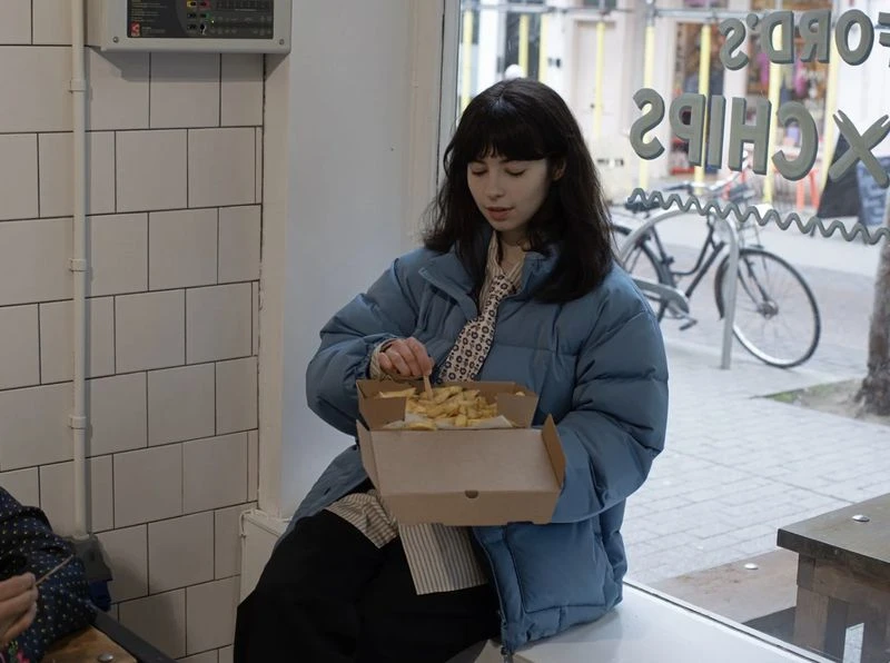 Young woman with dark hair sits indoors eating chips from a cardboard container, wearing a blue puffer jacket and patterned tie.