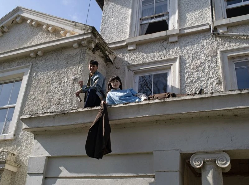 Two people sit on the ledge of a building. An adult male holds a paint roller and a child sits in a blue shirt. A jacket hangs from the ledge.