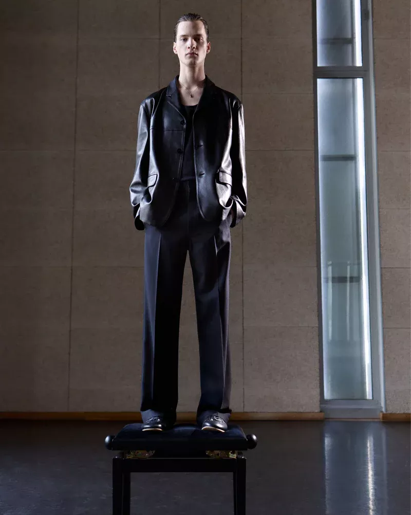 Man in a dark suit and black leather jacket stands on a small platform indoors with modern architectural details. Fashion editorial.