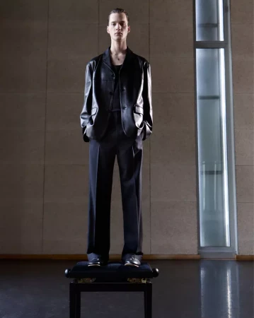 Man in a dark suit and black leather jacket stands on a small platform indoors with modern architectural details. Fashion editorial.