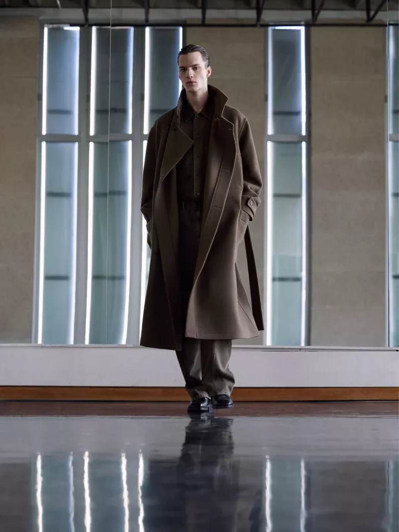 Fashion shot of a male model in a brown coat and trousers walking on a reflective floor with a light wall in the background.