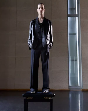 Model standing on a stool wearing a black leather jacket and trousers against a tiled wall