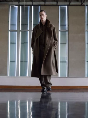 Model wearing a long brown belted coat standing in front of tall vertical windows with a polished reflective floor.