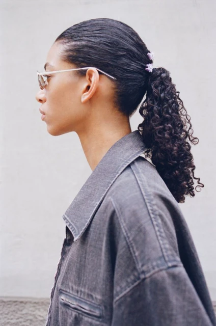 Profile of a young adult female model with dark curly hair in a ponytail, wearing a grey denim jacket and silver framed sunglasses.