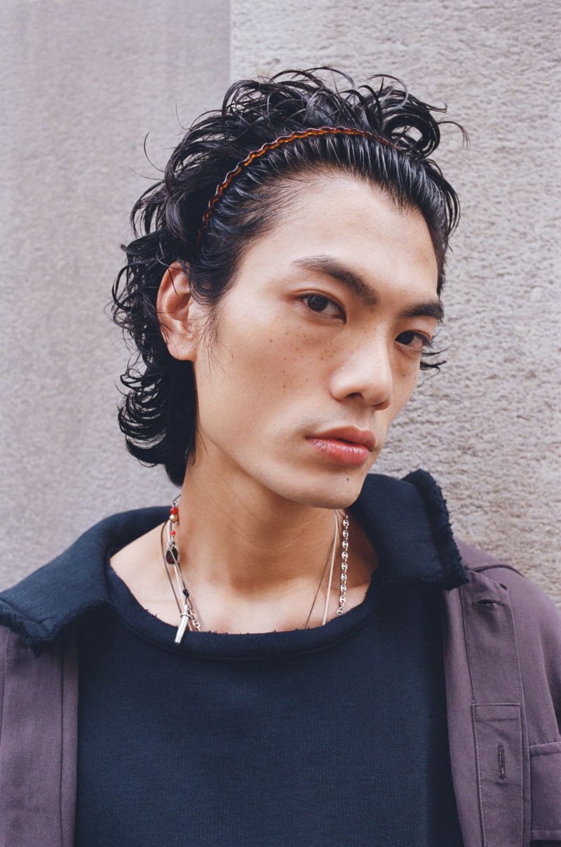 Portrait of an adult male model with dark curly hair and a brown headband, posing in a dark shirt and jacket against a gray wall.