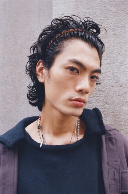 Portrait of an adult male model with dark curly hair and a brown headband, posing in a dark shirt and jacket against a gray wall.