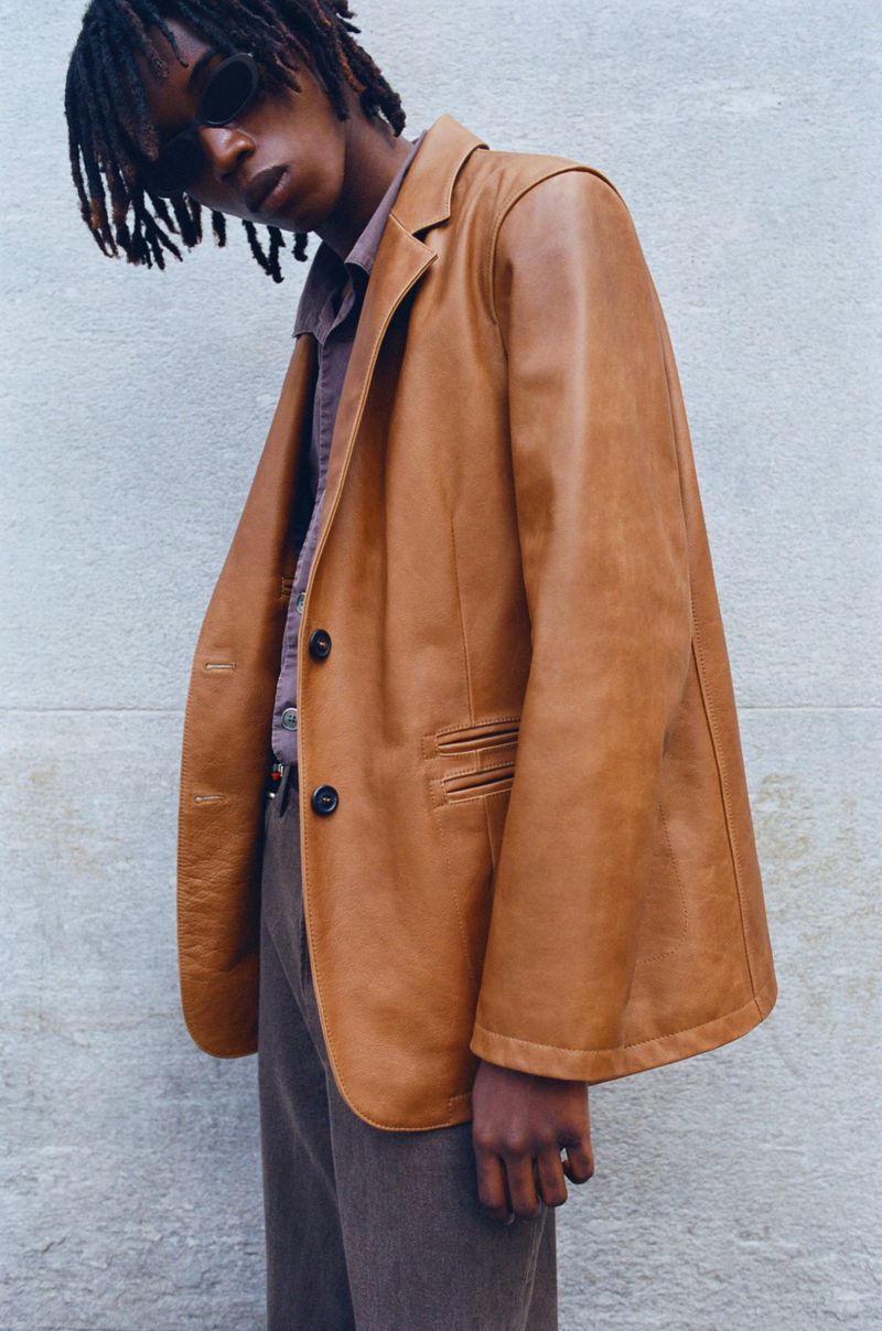 Portrait of a man wearing a tan leather blazer, brown shirt, and sunglasses against a plain stone wall background