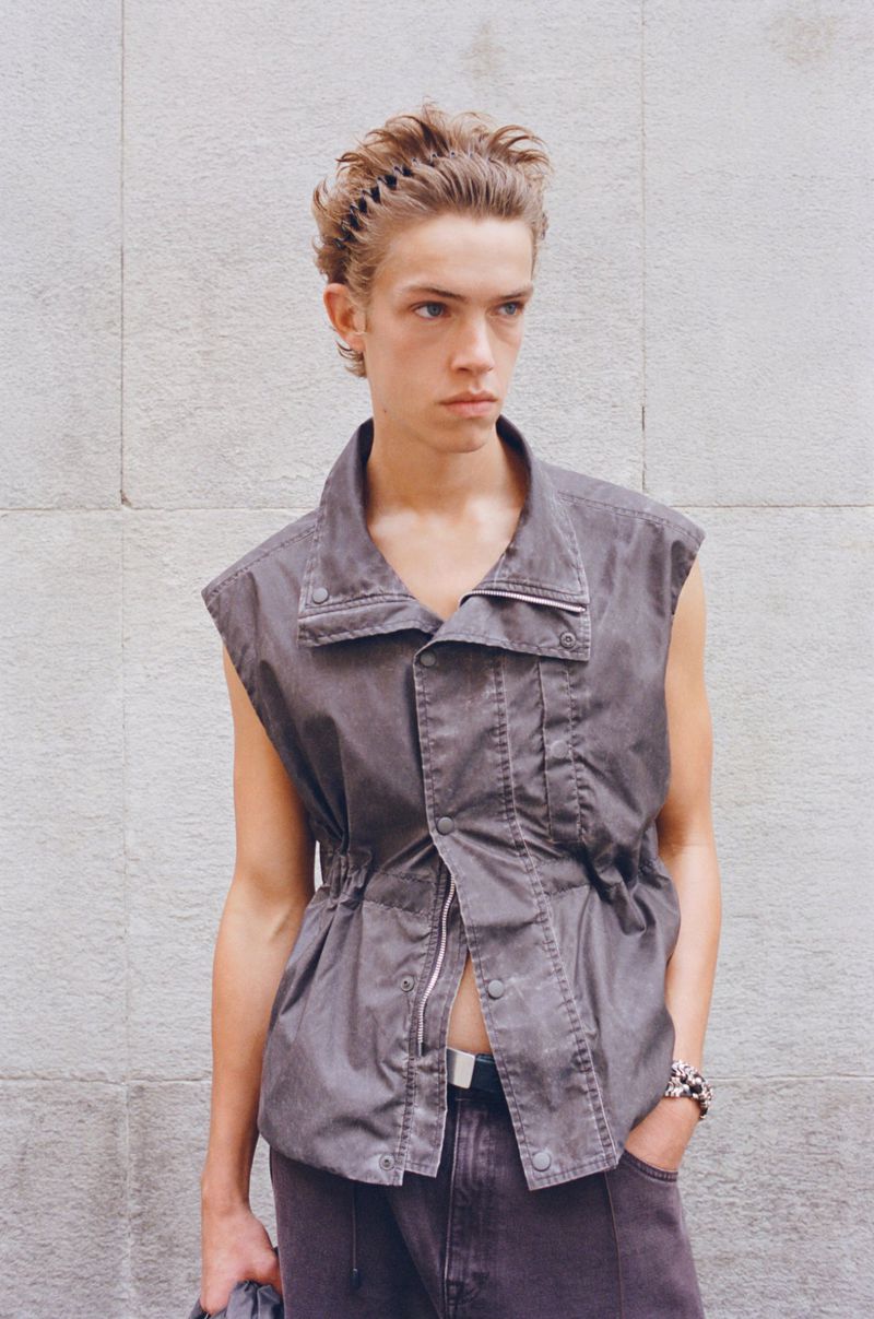 A young male fashion model poses in a sleeveless button-down top and jeans in front of a grey wall, looking off-camera with a neutral expression.
