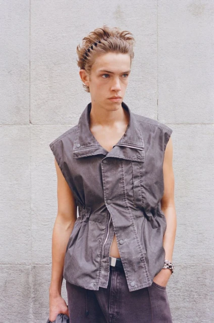 A young male fashion model poses in a sleeveless button-down top and jeans in front of a grey wall, looking off-camera with a neutral expression.