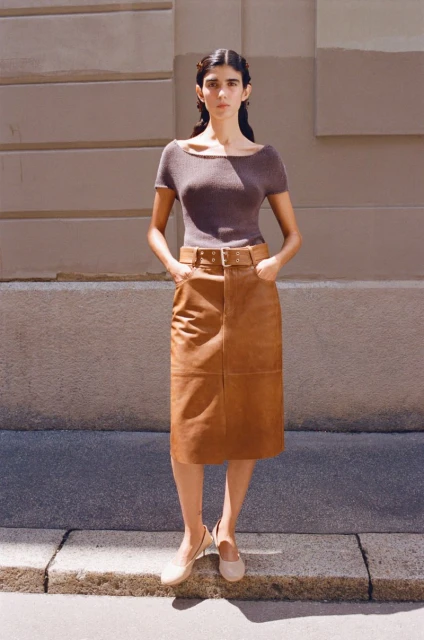 A woman stands outdoors wearing a brown short-sleeved shirt, brown leather skirt, and cream-colored shoes against a neutral wall.