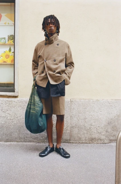 Adult male model in a tan jacket and shorts poses on a sidewalk next to a light-yellow building with a window and a gray sidewalk.