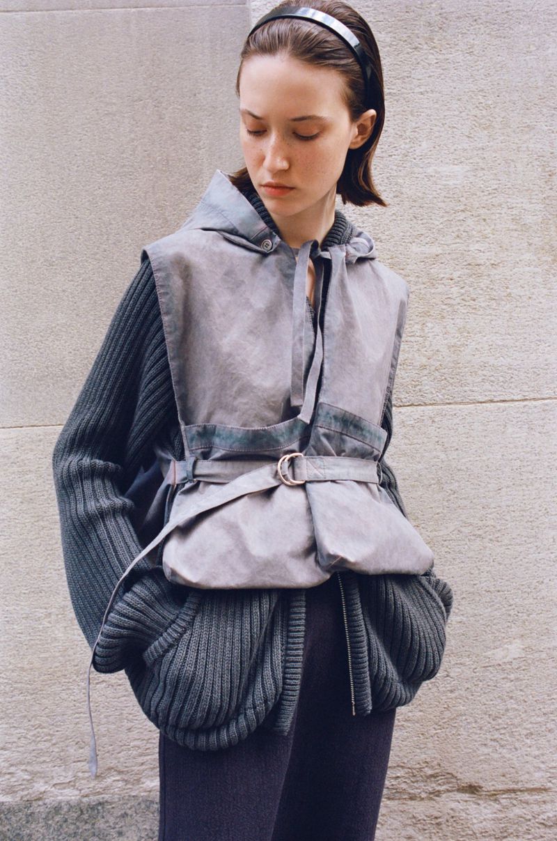 A young woman poses in a gray hooded vest with a belt over a dark green sweater, against a textured concrete wall with soft, diffused light.