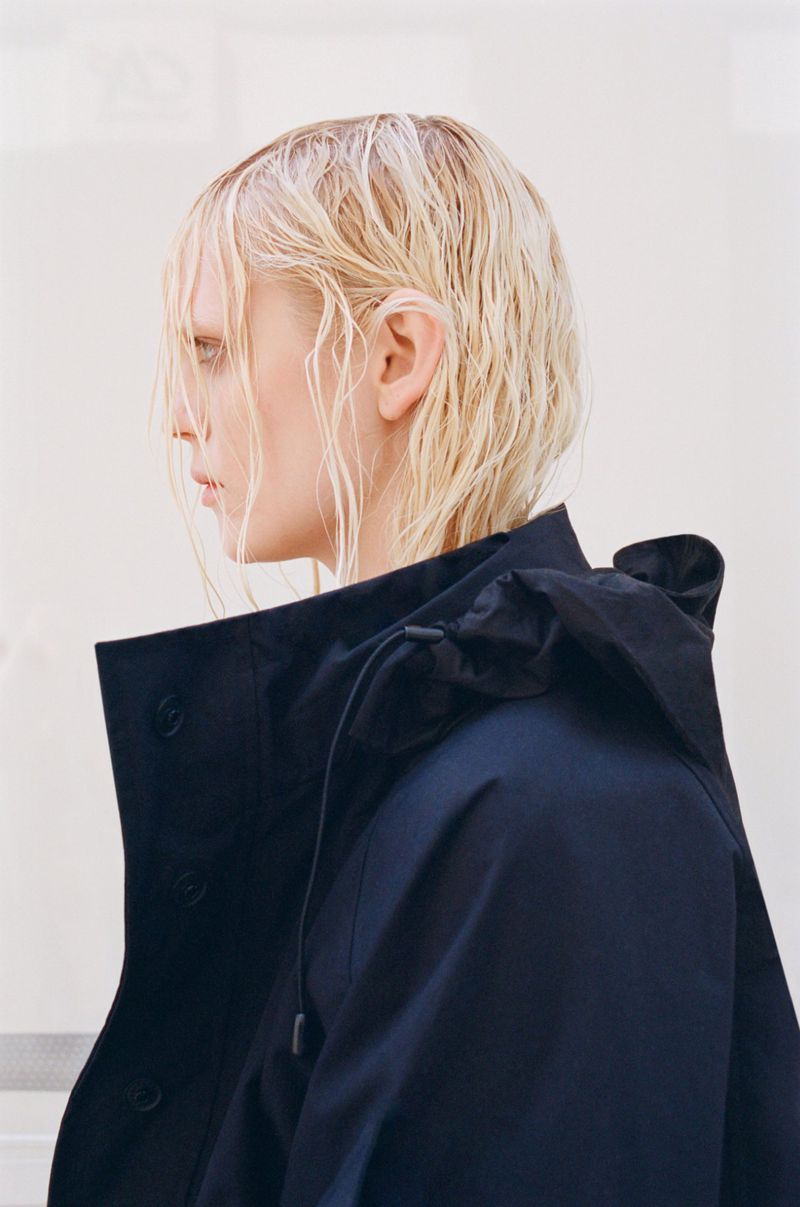 Profile view of a blonde adult woman wearing a dark navy jacket, set against a pale white background. The jacket has a hood and button detail.