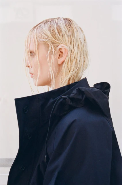 Profile view of a blonde adult woman wearing a dark navy jacket, set against a pale white background. The jacket has a hood and button detail.