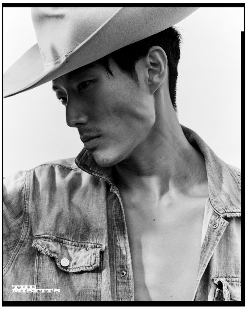 Black and white portrait of an Asian man wearing a wide-brim cowboy hat and unbuttoned denim shirt looking downward.