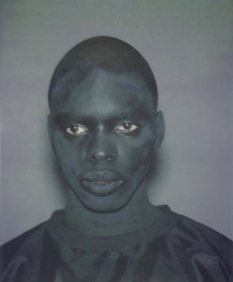 Close-up portrait of a person with dark skin and a dark shirt, set against a muted grey background, focusing on facial features.