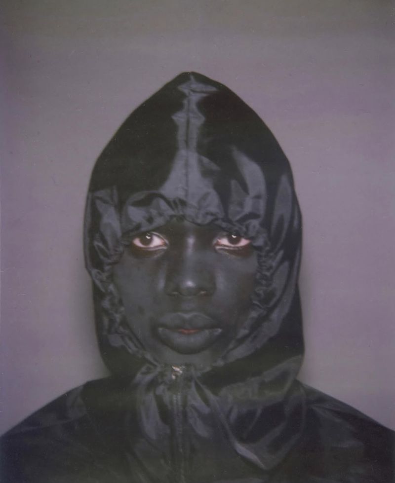 Portrait of a person with dark skin wearing a shiny dark hooded jacket, standing against a grey backdrop, captured in a medium studio shot.