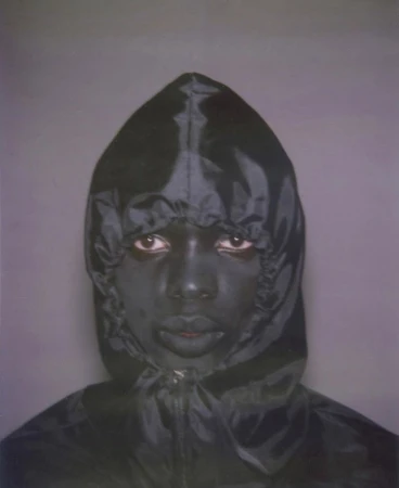 Portrait of a person with dark skin wearing a shiny dark hooded jacket, standing against a grey backdrop, captured in a medium studio shot.