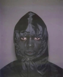Model Christ Eshiba Emir wearing a black hooded jacket zipped up to the chin against a grey background.