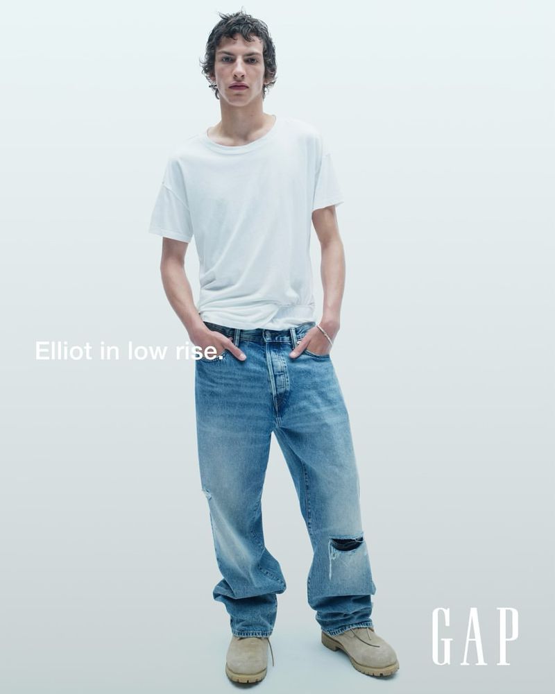 A young adult male posing in a casual outfit consisting of a white t-shirt, light-wash ripped jeans, and tan shoes against a bright white backdrop.