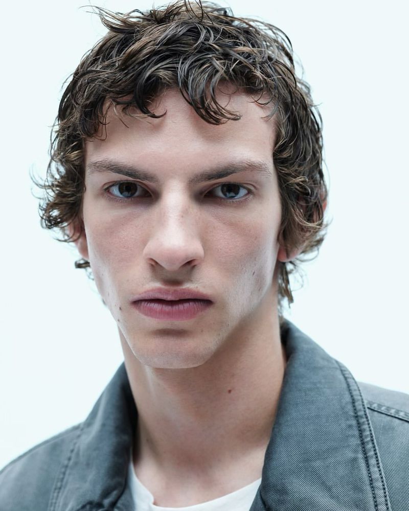Close-up studio shot of a young adult male with curly brown hair, wearing a gray jacket and white t-shirt, against a white background.