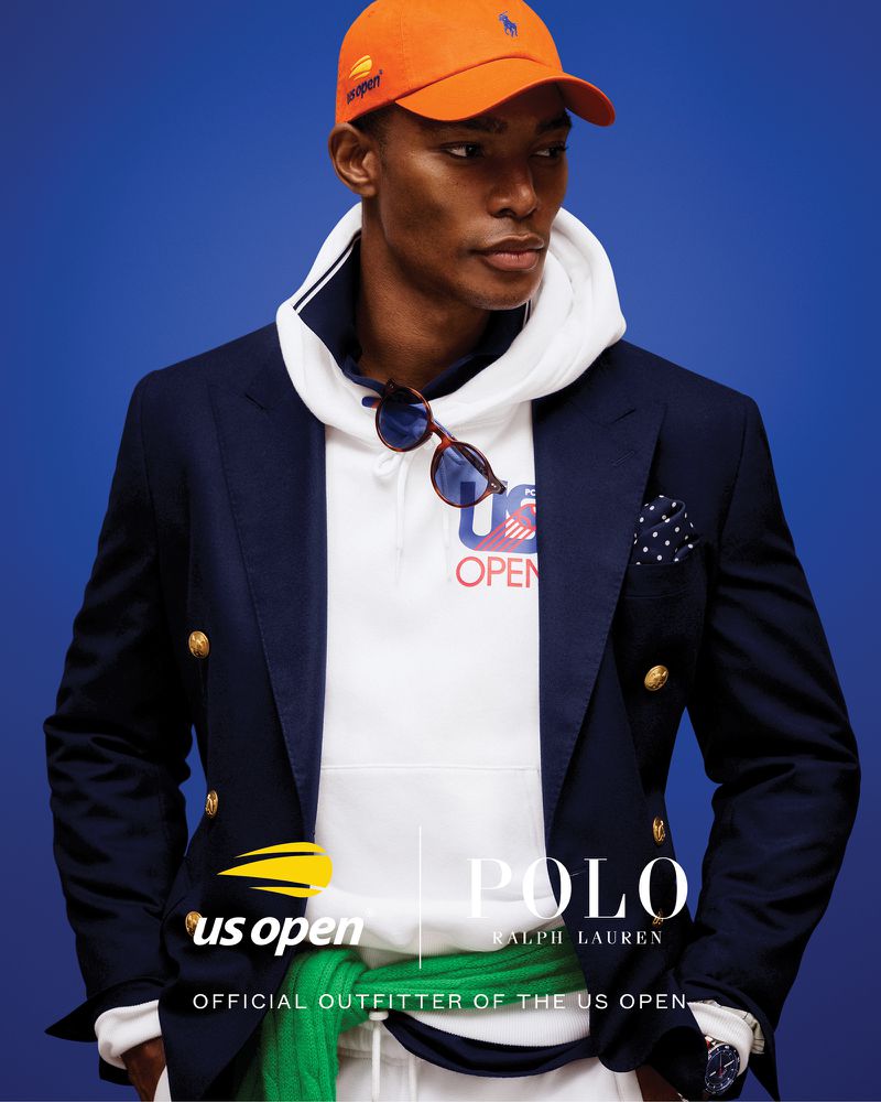 A dark-skinned male model wears a blue blazer, white hoodie and pants, with an orange baseball cap, posing against a blue background.