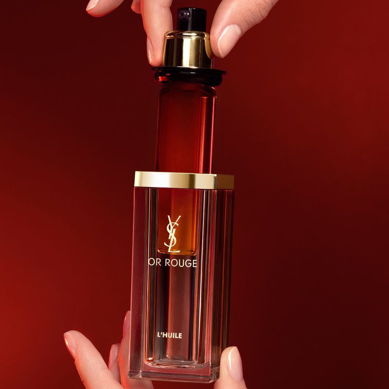 Close-up studio shot of YSL Or Rouge L'Huile bottle held by two hands against a red background, highlighting the product and its packaging.
