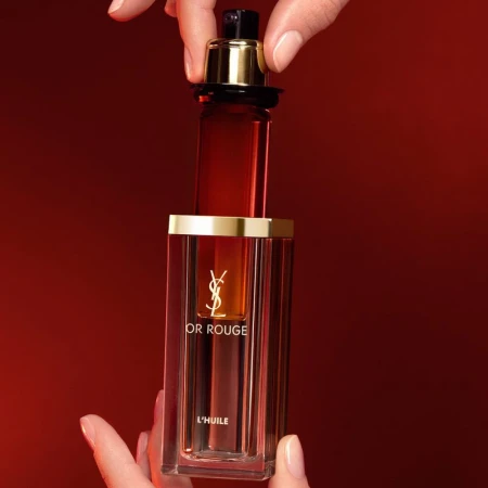 Close-up studio shot of YSL Or Rouge L'Huile bottle held by two hands against a red background, highlighting the product and its packaging.