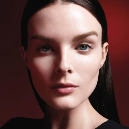 Close-up beauty shot of an adult female model with fair skin, black hair, blue eyes, and neutral expression against a red background.