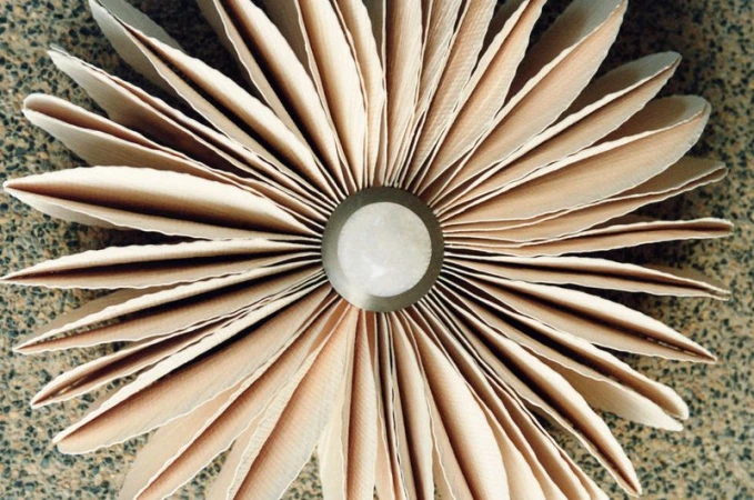 Overhead shot of an abstract floral arrangement crafted from layers of tan and cream-colored paper, radiating from a central gray core.