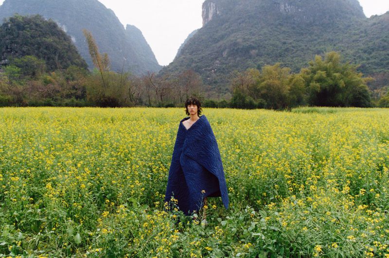 Adult stands in a field of yellow flowers wearing a blue quilt with misty mountains in the background on an overcast day.