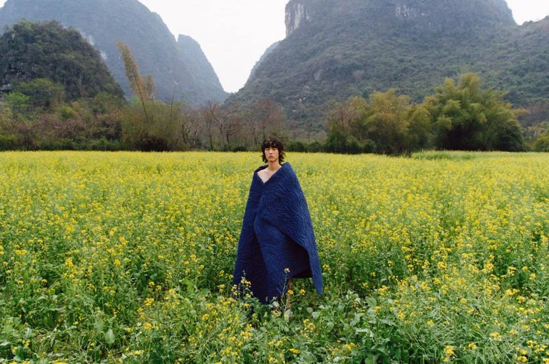 Adult stands in a field of yellow flowers wearing a blue quilt with misty mountains in the background on an overcast day.