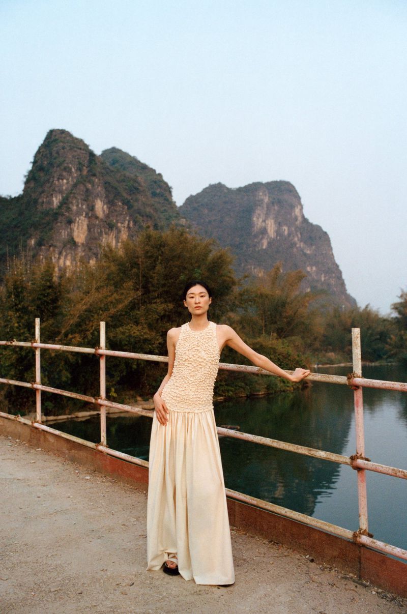 Woman in a beige dress poses on a bridge with mountains and river in the background, creating an editorial fashion image.